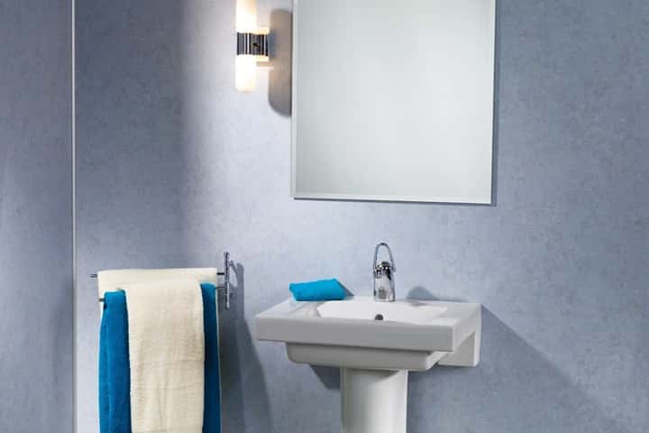 Witte staande wastafel tegen een blauwgrijze wand met wandlamp, spiegel en blauwe handdoeken.