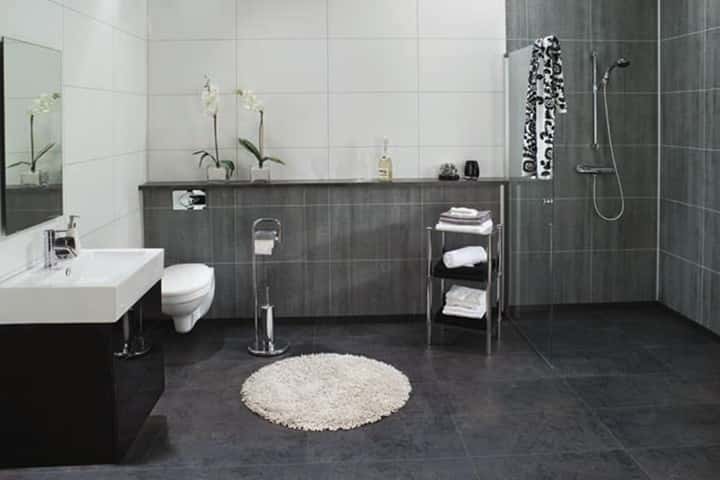 Badkamer met donkergrijze leisteenlook-wandpanelen, een witte wastafel met wandkraan en een ronde witte badmat.