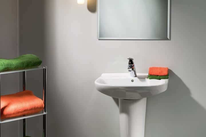 Witte staande wastafel tegen een gladde lichtgrijze wand, met oranje en groene handdoeken op een chromen rek.
